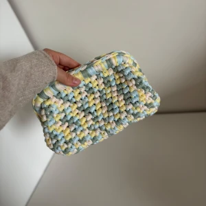 Pastellfärgad virkad necessär från Design.by.bela - Superfin handgjord axelväska i pastelliga toner av blått, gult och vitt. Väskan är virkad med tjockt garn och har en mjuk, fluffig känsla. Perfekt för att addera färg till din outfit! Gå gärna in på Instagram @design.by.bela för andra necessärer och väskor. 