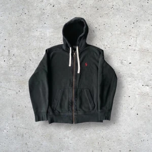 Svart hoodie från Polo Ralph Lauren - Säljer en svart hoodie från Polo Ralph Lauren med dragkedja och vit snörning i huvan. Klassisk röd logga broderad på bröstet. Perfekt för en avslappnad stil och enkel att matcha med det mesta.