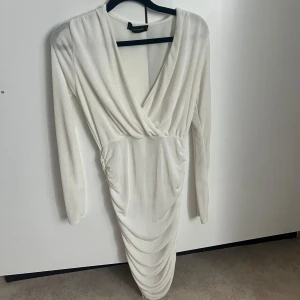 Vit glittrig omlottklänning från NA-KD - Säljer en vit glittrig omlottklänning från NA-KD med lång ärm och draperad kjol. Klänningen har v-ringning och är figurnära med snyggt fall. Perfekt för dig som vill sticka ut med en elegant look.