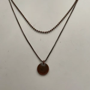Bronsfärgat halsband med rund berlock - Säljer ett lite slitet men snyggt dubbelt halsband i bronsfärg. Det ena lagret har små kulor och det andra en tunn kedja med en rund, slät berlock. Perfekt för att ge din outfit en cool touch. 