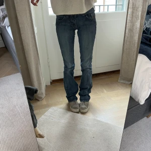Blå bootcut jeans från Cowgirl Tuff Co - Säljer ett par klassiska blå bootcut jeans från Cowgirl Tuff Co. Jeansen har normal passform, snygga detaljer på bakfickorna och en något sliten look. Storlek 34 och väldigt långa i benen, i fint skick. Jag är 169 cm lång, och på bilderna är mitt innerbensmått ca 82,5 cm och midjemått (platt mätt på mig) ca 31 cm.