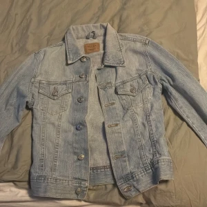 Ljusblå jeansjacka från Rocky - Säljer en klassisk ljusblå jeansjacka från Rocky med knappar framtill och två bröstfickor med lock. Jackan har en tidlös design med krage och justerbara knappar i midjan. Perfekt att slänga över en t-shirt för en avslappnad look.