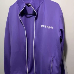 Lila track jacket från Palm Angels - Säljer en lila track jacket från Palm Angels med vit logga på bröstet och vita ränder längs ärmarna. Jackan har dragkedja framtill och ståkrage, samt fickor med dragkedja. Perfekt för en sportig och trendig look. Pris kan diskuteras vid snabb affär!
