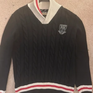 Svart kabelstickad tröja från Morris - Svart kabelstickad tröja från Morris med vit och röd ribbad krage samt muddar. Tröjan har lång ärm, v-ringad krage och patchar med Morris-logga och brittisk flagga. Jättefint skick, skriv för mer info eller bilder!