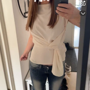 Beige omlottopp med knytband - Säljer en stilren beige omlottopp med knytband i midjan. Toppen har en rund halsringning och är ärmlös, vilket gör den perfekt för varmare dagar eller lager på lager. Passar till både jeans och kjol. Använd 2 gånger max, som ny