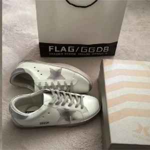 Vita sneakers från Golden Goose med silverdetaljer - Säljer ett par vita sneakers från Golden Goose med silverfärgad stjärna på sidan och silverdetaljer vid hälen. Skorna har grå snörning och en platt sula. Klassisk och stilren design med märkets logga på hälen.