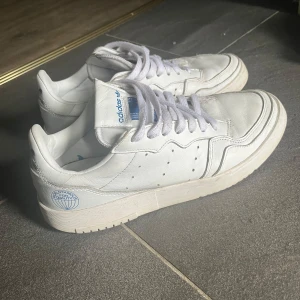 Vita Adidas sneakers med blå logga - Säljer ett par klassiska vita Adidas sneakers med blå logga på hälen och tungan. Skorna har snörning, rund tå och platt sula. Perfekta för en stilren och sportig look.
