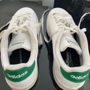 Vita Adidas Originals Advantage Base M knappt använda - Säljer ett par klassiska vita Adidas sneakers med gröna detaljer på hälen och logga. Skorna har snörning, rund tå och platt sula. Perfekta för en clean och sportig stil. Adidas logga syns tydligt både bak och på plösen.                                  Original pris: 930kr från zalando