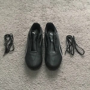 Svarta fotbollsskor från Puma - Säljer ett par svarta fotbollsskor från Puma med vita detaljer och klassisk design. Skorna har rund tå, platt sula och medföljande svarta skosnören. Perfekta för dig som vill ha stilrena och funktionella skor på planen.
