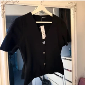 Svart blus från Gina - Snygg svart blus från Gina Tricot med v-ringning och korta ärmar. Blusen har tre dekorativa knappar framtill som ger en elegant touch. Perfekt att styla till olika outfits.