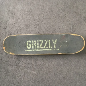 Skateboard med Grizzly griptape och grafiskt mönster - Säljer en skateboard med svart Grizzly griptape och grafiskt mönster i grönt och gult på undersidan. Brädan har svarta trucks och slitna, beige och svarta spit fire hjul. Passar dig som gillar street och skatekultur.