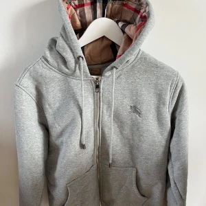 Grå  Burberry hoodie med dragkedja - Säljer en grå hoodie från Burberry med dragkedja och klassiskt rutigt foder i huvan. Storlek M passar också storlek S. 