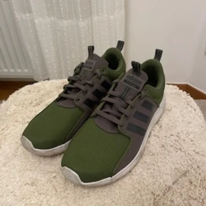 Adidas sneakers i grönt och grått - Säljer ett par Adidas sneakers i grönt med grå detaljer och svarta ränder på sidorna. Skorna har snörning och vit platt sula. Perfekta för vardagsbruk och träning.