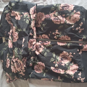 Blommig jacka från KappAhl - Säljer en svart jacka från KappAhl med rosa blommigt mönster och gröna blad. Jackan har dragkedja framtill och tryckknappar på fickorna. Perfekt för dig som gillar färg och mönster!