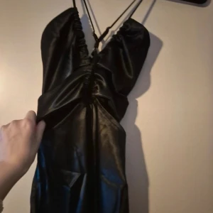 Svart långklänning med slits och cut-out detaljer - Säljer en svart långklänning med smala axelband, cut-out detaljer framtill och hög slits på ena sidan. Klänningen har en glansig finish och en snygg omlottkänsla över bysten. Inte använd 