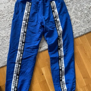 Blå Adidas träningsbyxor med logga - Snygga blå träningsbyxor från Adidas med svarta och vita loggor längs båda benen. Byxorna har resår i midjan. perfekta för sport eller chill. Klassisk streetstil med tydlig Adidas-branding.
