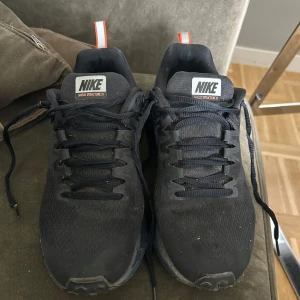 Svarta Nike Shield Structure 21 sneakers - Säljer ett par svarta Nike Shield Structure 21 sneakers med orange och reflekterande detaljer. Skorna har snörning, rund tå och slitstark sula med hexagonmönster. Perfekta för löpning eller vardagsbruk, vattenavvisande och håller värmen.