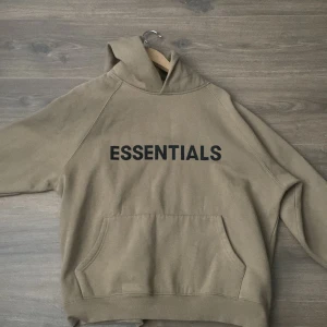 Beige Essentials hoodie - Säljer en beige hoodie från Essentials med svart text på bröstet. Tröjan har huva, magficka och ribbade muddar. Perfekt för en avslappnad och stilren look. Storlek S.