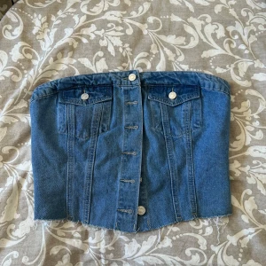 Jeans korsett-topp med knappar - Säljer en snygg korsett-topp i blå jeans med knappar framtill och fickdetaljer. Toppen har smockad rygg för bättre passform och råa kanter nedtill för en trendig look. Perfekt att styla till olika outfits!