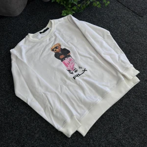 Vit sweatshirt från Polo Ralph Lauren - Vit sweatshirt från Polo Ralph Lauren med ett stort tryck av en björn på framsidan. Tröjan har rund halsringning och RLX-tryck under motivet. Tillverkad i 100% bomull för en mjuk och bekväm känsla.