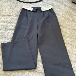 Grå kostymbyxor med vit midjedetalj - Snygga grå kostymbyxor med en unik vit detalj i midjan. Byxorna har raka ben och pressveck framtill, vilket ger en stilren och modern look. Perfekta för dig som vill sticka ut lite extra.