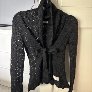 Odd Molly stickad kofta med knytband - Säljer en svart stickad kofta från Odd Molly med knytband framtill. Koftan har lång ärm och en mjuk, luftig känsla. Perfekt att slänga över en topp för en snygg lager-på-lager-look.