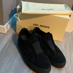 Svarta sneakers från Axel Arigato - Säljer ett par svarta sneakers från Axel Arigato i mocka med minimalistisk design. Skorna har rund tå, platt sula och saknar synlig snörning för en clean look. Perfekta för dig som gillar stilrena och moderna skor.