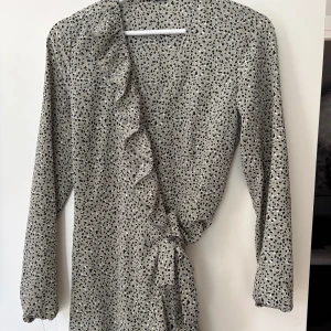 Prickig omlottklänning med volang - Säljer en snygg omlottklänning med svart prickigt mönster på ljusgrön botten. Klänningen har långa ärmar och volangdetalj längs omlottkanten. Använd fåtal gånger.