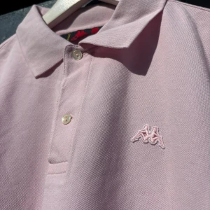 Ljusrosa pikétröja från Kappa - Säljer en ljusrosa pikétröja från Kappa med klassisk krage och två knappar framtill. Tröjan har korta ärmar och Kappas logga broderad på bröstet. Perfekt för en avslappnad och sportig stil.