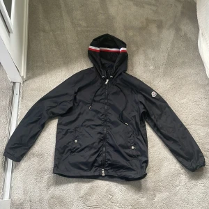 Svart vindjacka med huva från Moncler - Säljer en svart vindjacka från Moncler med huva och dragkedja. Jackan har en röd, vit och blå rand på huvan och två fickor framtill med knappar. Perfekt för blåsiga dagar och enkel att matcha.