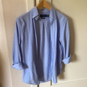 Ljusblå skjorta från Polo Ralph Lauren - Klassisk ljusblå skjorta från Polo Ralph Lauren i modellen Knit Oxford. Skjortan har button-down krage och långa ärmar, perfekt för en stilren look. Tillverkad i bomull för en bekväm känsla.