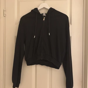 Svart croppad hoodie med dragkedja - Säljer en svart kort hoodie med dragkedja framtill och huva med snören. Den har långa ärmar och resår i midjan.