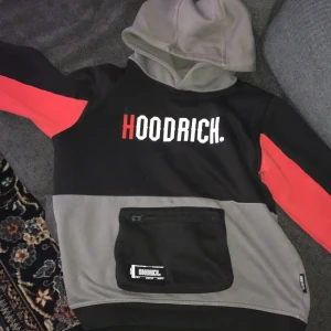 Hoodrich hoodie med röd detalj - Säljer en hoodie från Hoodrich i svart, grått och rött. Tröjan har stor magficka med dragkedja, tryckt logga fram och bak samt huva. Perfekt för dig som gillar streetwear och vill sticka ut med snygga färgblock.