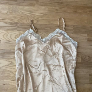 Guldig silkelinne med spetsdetaljer - Säljer en guldig pyjamas linne i satin med tunna axelband och v-ringning. Toppen har fina spetsdetaljer längs urringningen och är tillverkad i ett glansigt material som ger en lyxig känsla.