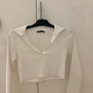 Vit croppad långärmad topp från Zara - Säljer en stilren vit croppad topp från Zara med lång ärm och krage. Toppen är ribbad och har en djup v-ringning framtill. Perfekt att matcha med jeans eller kjol för en trendig look.