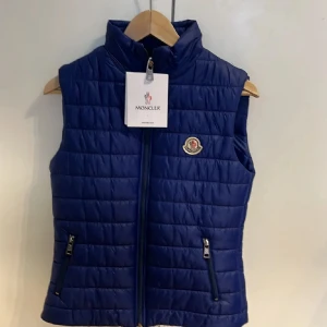  Moncler väst äkta* - Snygg blå dunväst från Moncler med ståkrage och dragkedja framtill. Västen har två fickor med dragkedja och Moncler-logga på bröstet. Perfekt för lager-på-lager och ger en sportig look.