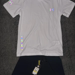 Vit t-shirt från Under Armour - Säljer en vit t-shirt från Under Armour med färgglada loggor och detaljer. T-shirten har rund halsringning och korta ärmar, perfekt för träning eller sportiga aktiviteter.