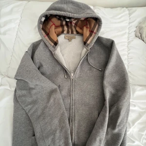 Grå hoodie från Burberry med klassiskt rutigt foder - Säljer en grå hoodie från Burberry med dragkedja och känguruficka. Insidan av huvan har det ikoniska Burberry-rutiga fodret i beige, rött och svart. Perfekt för dig som vill ha en stilren och lyxig vardagströja.