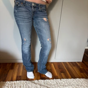 Blå jeans från True Religion med slitningar - Säljer ett par blå jeans från True Religion med raka ben och snygga slitningar framtill. Jeansen har dragkedjor på bakfickorna och klassiska detaljer. Perfekta för dig som gillar en avslappnad men trendig look.