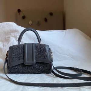 Mörkgrå axelväska i teddy-material - Säljer en mörkgrå axelväska från hm med handtag och justerbar axelrem. Väskan är gjord i mjukt teddy-material med detaljer i syntetläder. Magnetlås framtill och stilren design som sticker ut.