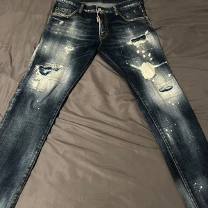 Dsquared2 jeans med slitningar och tryck - Mörkblå jeans från Dsquared2 med tydliga slitningar, hål och färgstänk på framsidan. Klassisk femficksmodell med gylf och knapp, samt broderad detalj och tryck bak. Snygga kontrastsömmar och en cool, edgy look.