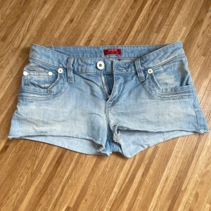 Lågmidjade ljusblå jeansshorts - Såå fina ljusblå jeansshorts från Raer med slitna detaljer och råa kanter. Klassisk femficksmodell med vita knappar och jättesnygga stenar på bakfickorna i vitt och blått. Skick enligt bilderna. Vet tyvärr inte storleken helt, står storlek 12 på dem, men skulle säga att de sitter som en xxs-xs, midjemåttet rakt över är ca 34 cm som visas på bilden 💗
