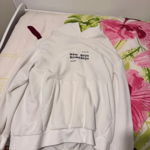 beige hoodie - Säljer en beige hoodie med svart texttryck på bröstet där det står 'New days Nowadays'. Hoodien har en stor magficka och huva. Perfekt för en avslappnad stil.