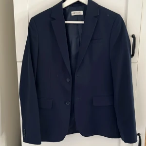 Mörkblå kavaj från H&M - Säljer en stilren mörkblå kavaj från H&M i klassisk modell. Kavajen har två knappar framtill, fickor med lock och är helfodrad med diskret prickigt innerfoder. Perfekt för bal eller festliga tillfällen.