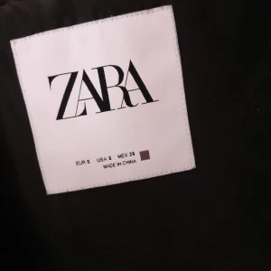 Svart quiltad jacka med huva från ZARA - Säljer en svart quiltad jacka från ZARA i storlek S. Jackan har huva, dragkedja framtill och två stora fickor. Perfekt för kyligare dagar och enkel att matcha med olika outfits.