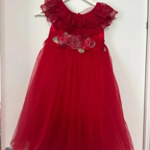 Röd långklänning med spets och blommor - Säljer en röd långklänning med volangkrage i spets och dekorativa blommor i midjan. Klänningen har en romantisk känsla med mycket tyll och spetsdetaljer upptill. Perfekt för speciella tillfällen. För barn runt 8-9 år gammal.
