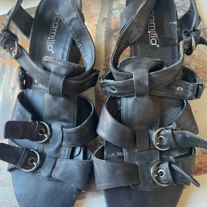 Svarta kilklack sandaler från Camilla - Säljer ett par svarta sandaler från Camilla med kilklack och flera remmar med spännen. Skorna har öppen tå och justerbara remmar för bra passform. Perfekta för varma dagar och ger en cool look till din outfit.