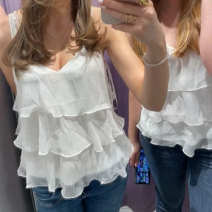 Vit volangtopp med smala axelband - Säljer en söt vit topp med flera lager volanger och smala axelband. Toppen har v-ringning och är tillverkad i ett lätt, luftigt material som ger en romantisk känsla. Perfekt för varma dagar eller när du vill känna dig extra fin.