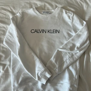 Vit sweatshirt från Calvin Klein Jeans - Vit sweatshirt från Calvin Klein Jeans med svart logotyptryck på bröstet. Klassisk rund halsringning och ribbade muddar vid ärmslut och nederkant. Tillverkad i 100% bomull för en mjuk och bekväm känsla.