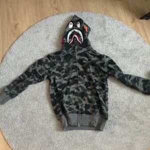 Camo hoodie från A Bathing Ape - Säljer en grå camouflagemönstrad hoodie från A Bathing Ape med dragkedja och ikonisk hajdesign på huvan. Tröjan har ribbade muddar och detaljerade broderier på huvan. Strl M men sitter lite mindre, jag är 182 (se preferens bild)